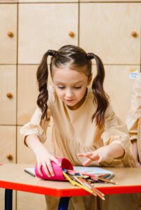 Young girl with ponytails organizing pencils from a pink holder on a school desk.