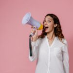 Woman in white shirt holding megaphone against a pink background, confidently speaking.
